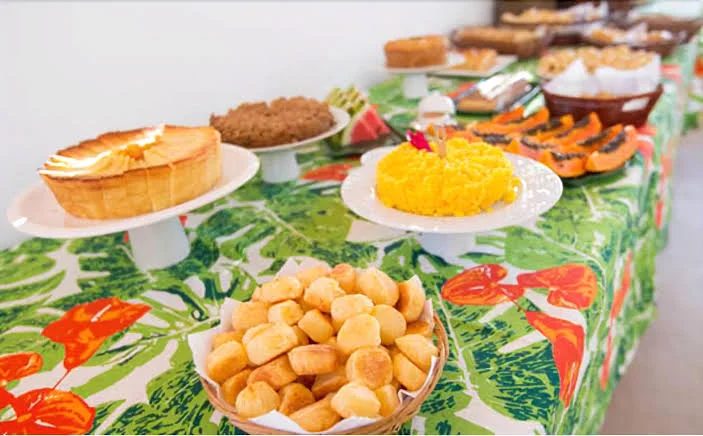 Mesa do café da manhã com bolos, pão de queijo, cuscuz e mamão sobre toalha estampada